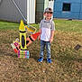 boy, building, cap, casual_clothing, child, crayons, daylight, educational, grass, jeans, kindergarten, leaves, outdoor, playful, school_bus, sidewalk, sign, smiling, sneakers, tree