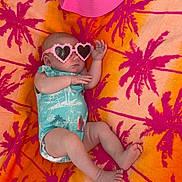 Nora is registered to the contest to win money with this photo: baby, infant, sunglasses, heart_shaped, towel, palm_tree_pattern, pink_hat, onesie, relaxing, summer, outdoor, colorful, cute, lying_down, barefoot, fashion, accessory, skin, head, hand