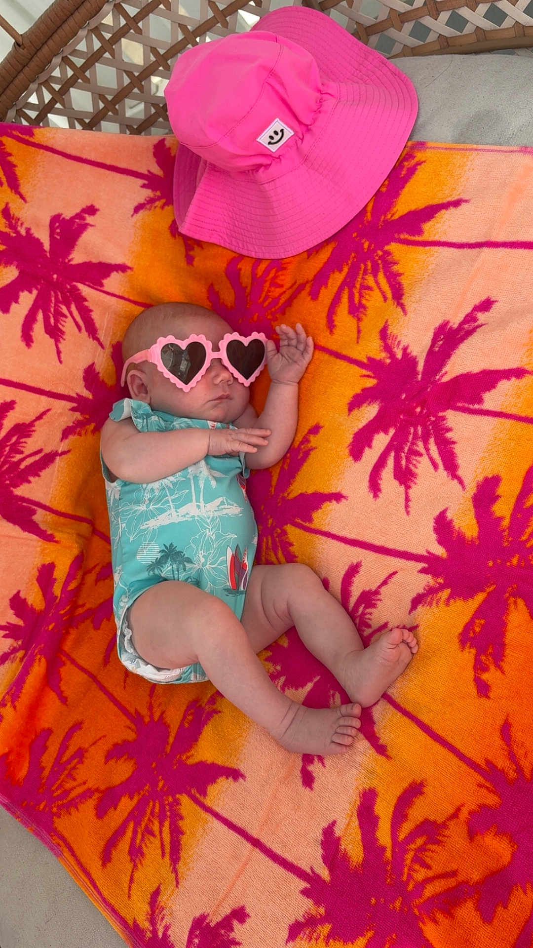 Nora is registered to the contest to win money with this photo: baby, infant, sunglasses, heart_shaped, towel, palm_tree_pattern, pink_hat, onesie, relaxing, summer, outdoor, colorful, cute, lying_down, barefoot, fashion, accessory, skin, head, hand