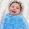 baby, child, flower_crown, smiling, blue_overalls, blanket, happy, portrait, infant, cute, face, headwear, soft_texture, buttoned_clothing, lying_down, cheerful, skin, eyes, hands, young_child