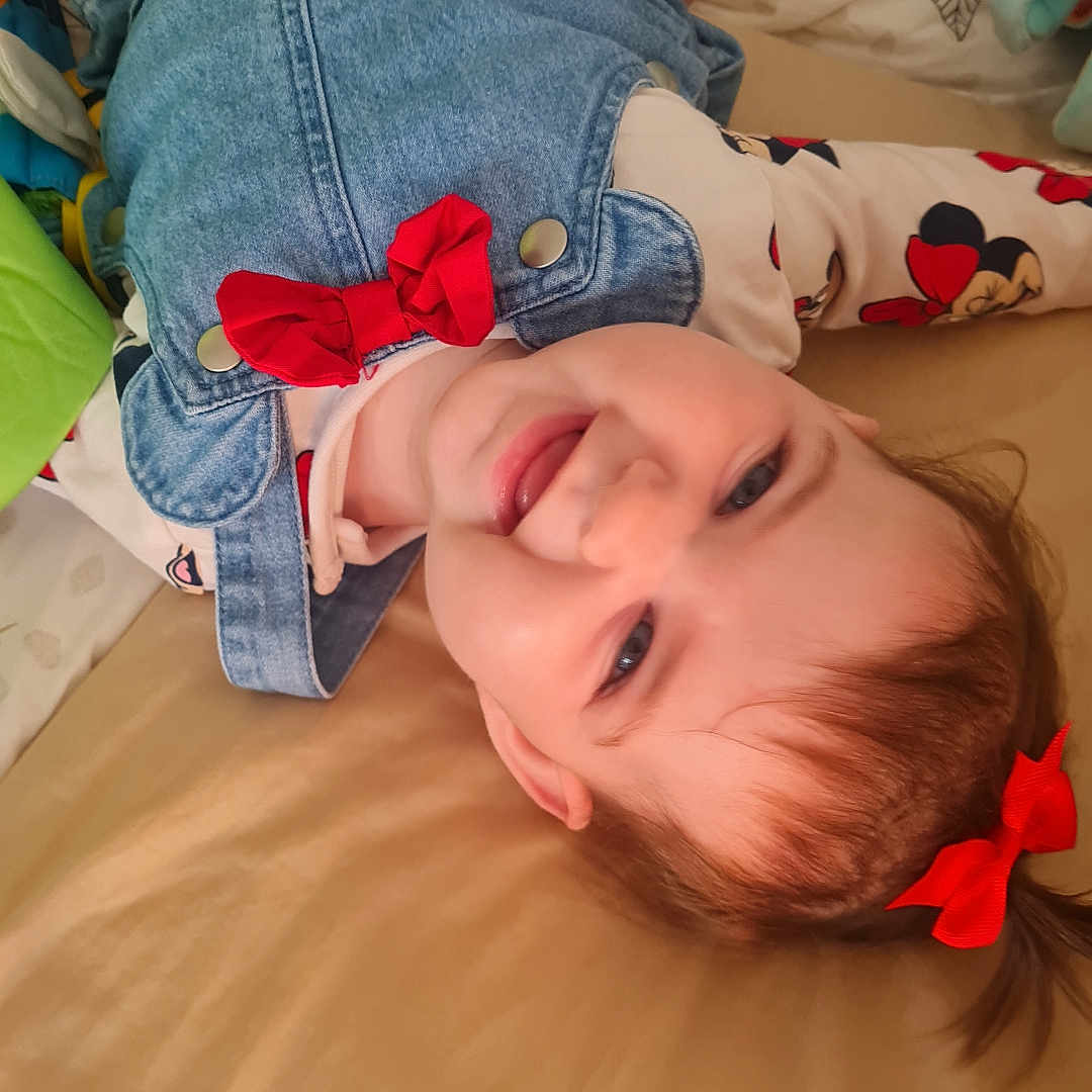 Luna a rejoint le concours — aidez-le/la à gagner de superbes lots ! baby, child, clothing, cute, denim_dress, face, hair_tie, happy, head, indoor, infant, lying_down, pattern, person, playful, red_bow, smiling, soft_surface, tongue_out, toy