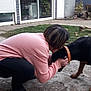 person, dog, outdoor, backyard, grass, patio, crouching, black_dog, orange_collar, casual_clothing, human_interaction, garden_tools, concrete, fence, house, window, door, plant, wheelbarrow, short_hair