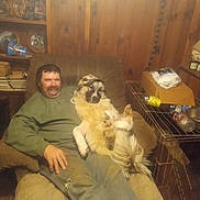 Buck is registered to the contest to win money with this photo: man, dog, chair, indoor, wooden_paneling, shelves, plates, books, cage, bottle, box, clothing, cap, relaxing, furniture, pet, smile, casual, seated, cozy