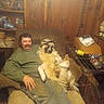 man, dog, chair, indoor, wooden_paneling, shelves, plates, books, cage, bottle, box, clothing, cap, relaxing, furniture, pet, smile, casual, seated, cozy