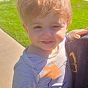 Adonis joined the competition — help win amazing prizes! toddler, child, boy, portrait, outdoor, sunlight, smiling, squinting, messy_hair, blond_hair, being_held, person, gray_shirt, long_sleeves, cartoon_shirt, grass, sidewalk, apartment_building, residential, daytime