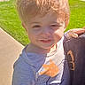 toddler, child, boy, portrait, outdoor, sunlight, smiling, squinting, messy_hair, blond_hair, being_held, person, gray_shirt, long_sleeves, cartoon_shirt, grass, sidewalk, apartment_building, residential, daytime