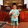 child, boy, thumbs_up, indoor, tshirt, plaid_shorts, carpet, microphone_stand, wood_paneling, bookshelf, smiling, face, short_hair, socks, cultural_setting, arabic_calligraphy, microphone, podium, red_line, carpeted_floor