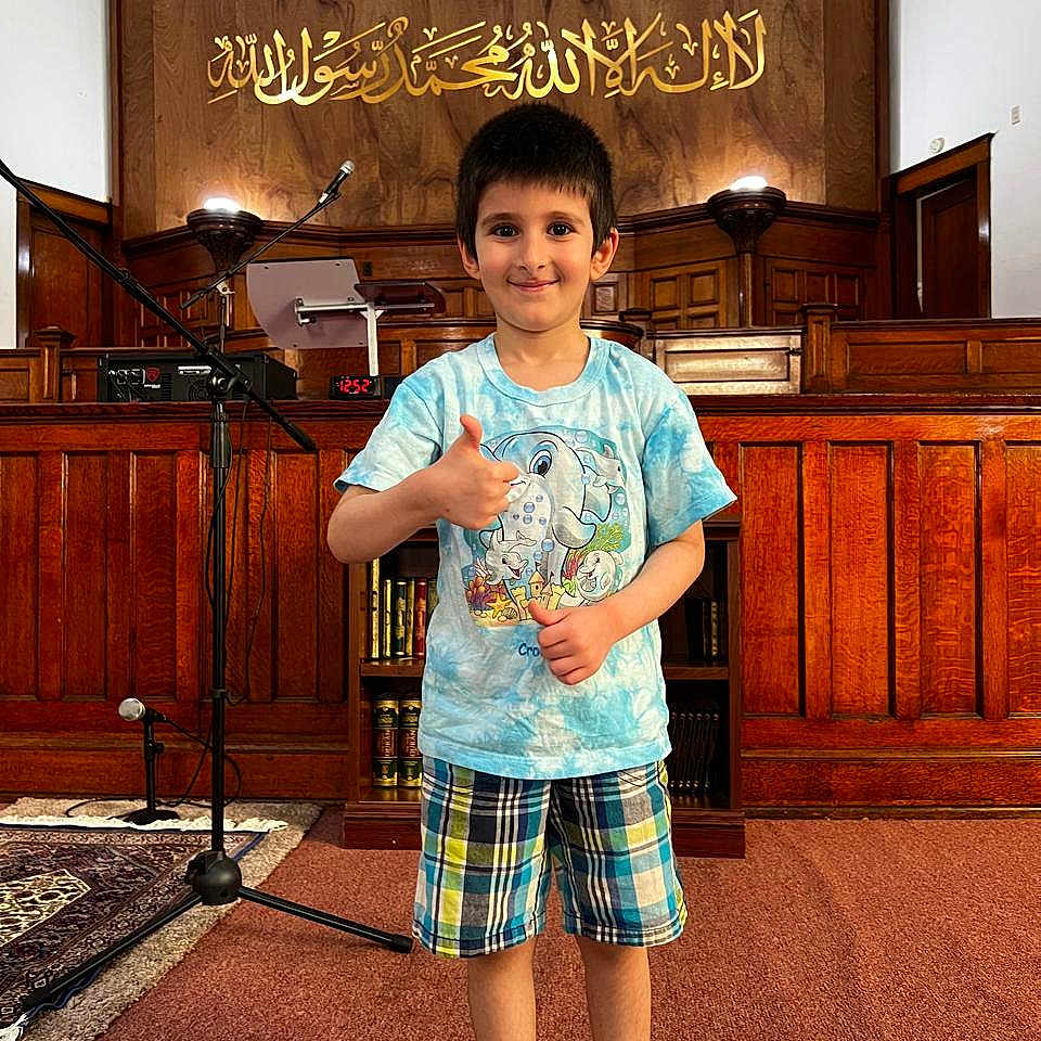 Essa is registered to the contest to win money with this photo: child, boy, thumbs_up, indoor, tshirt, plaid_shorts, carpet, microphone_stand, wood_paneling, bookshelf, smiling, face, short_hair, socks, cultural_setting, arabic_calligraphy, microphone, podium, red_line, carpeted_floor
