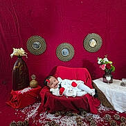 Alaia is registered to the contest to win money with this photo: baby, infant, smiling, red_backdrop, blanket, pinecones, vase, flowers, decorative_mirror, couch, holiday_scene, props, snow_flakes, bow, mittens, onesie, portrait, indoor, table, glass_jar