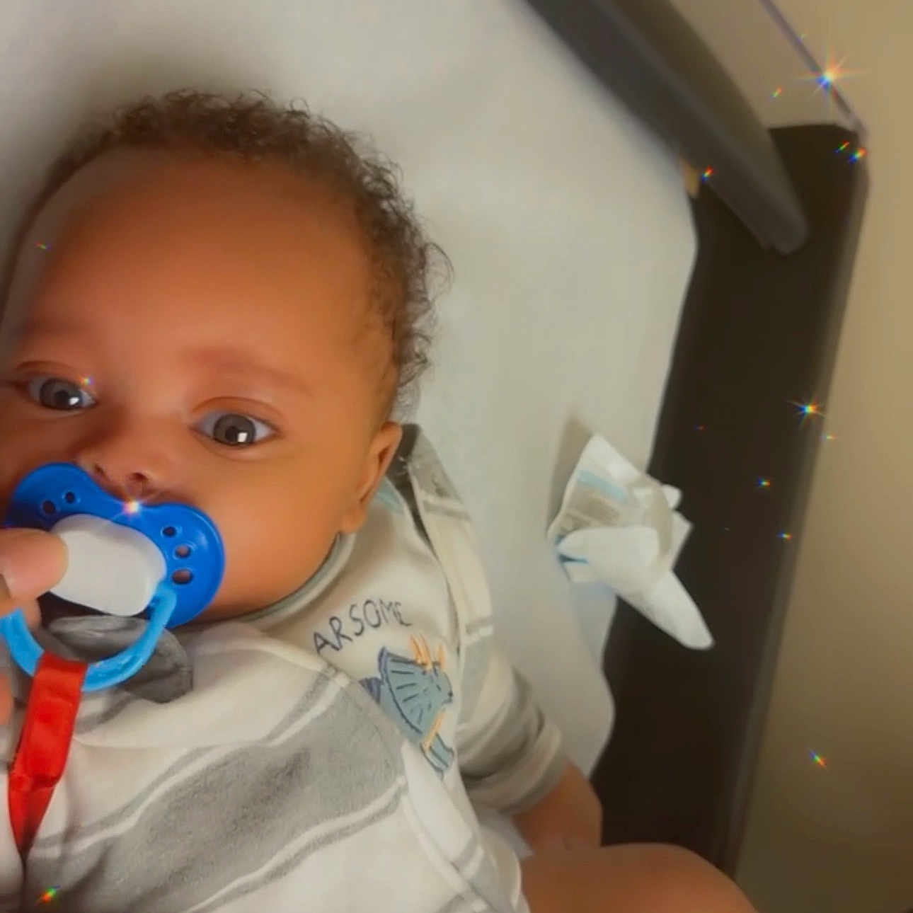 Dre'Vion is registered to the contest to win money with this photo: baby, pacifier, infant, medical_table, curly_hair, blue_pacifier, onesie, child, cute, lying_down, indoor, person, skin, face, hand, clothing, healthcare, examination, sparkle, closeup