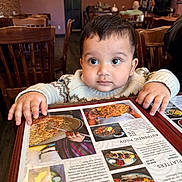 Azlan joined the competition — help win amazing prizes! baby, child, menu, restaurant, table, wooden_chair, curious, hands, clothing, indoor, food_photos, menu_cover, person, cute, face, eyes, sweater, seated, dining, portrait