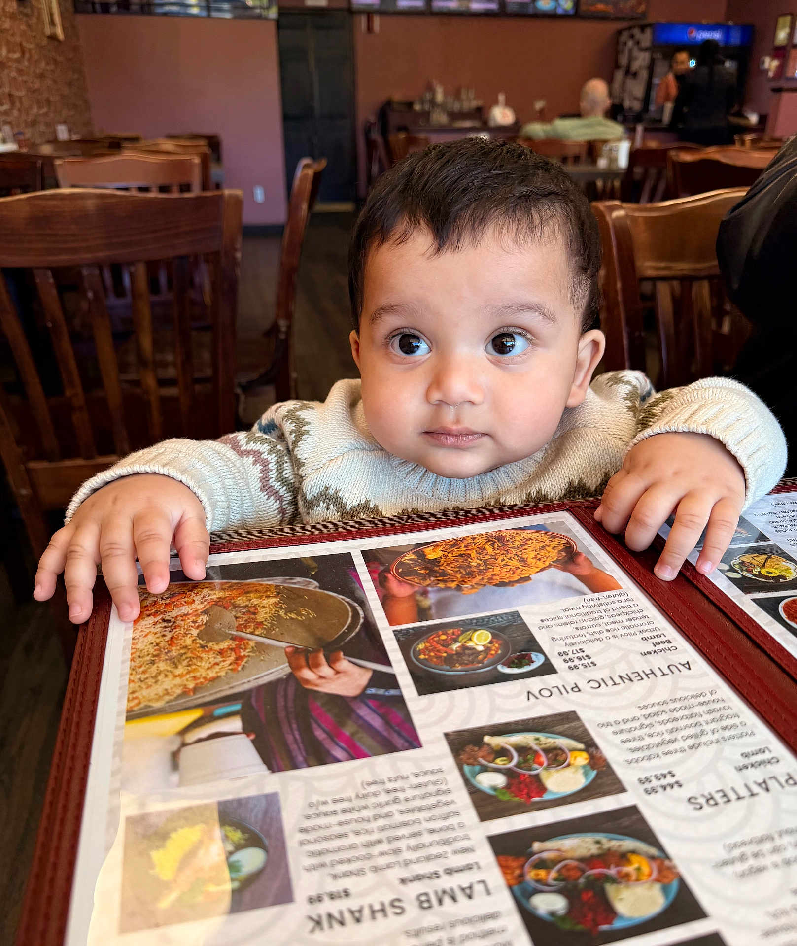 Azlan joined the competition — help win amazing prizes! baby, child, menu, restaurant, table, wooden_chair, curious, hands, clothing, indoor, food_photos, menu_cover, person, cute, face, eyes, sweater, seated, dining, portrait