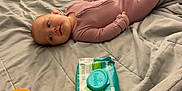 Aurora is registered to the contest to win money with this photo: baby, infant, bed, blanket, onesie, pampers, wipes, container, yellow_lid, fabric, sleeping_area, soft, cute, lying_down, indoor, person, child, room, curious, comfort