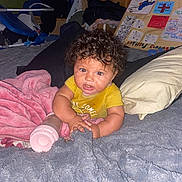 Ariyah joined the competition — help win amazing prizes! baby, child, curly_hair, yellow_shirt, blanket, pillow, bottle, bed, indoor, cluttered_background, poster, soft_texture, cozy, lying_down, infant, cute, expression, hands, face, room