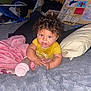 baby, child, curly_hair, yellow_shirt, blanket, pillow, bottle, bed, indoor, cluttered_background, poster, soft_texture, cozy, lying_down, infant, cute, expression, hands, face, room