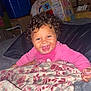 Ariyah joined the competition — help win amazing prizes! baby, curly_hair, smiling, blanket, bed, indoor, happy, child, person, pink_clothing, face, cheerful, portrait, infant, cozy, fabric, room, furniture, soft, wrapped