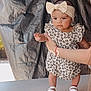 baby, child, girl, headband, bow, dress, floral_pattern, hand, supporting_hand, table, footwear, sandals, skin, face, expression, indoor, fabric_background, portrait, standing, person