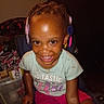 braids, candid_photo, casual_clothing, chair, child, colorful_clips, dark_skin, flash_photography, furniture, girl, green_shirt, hair_accessories, happy, home, indoors, pink_shorts, portrait, sitting, smiling, teeth