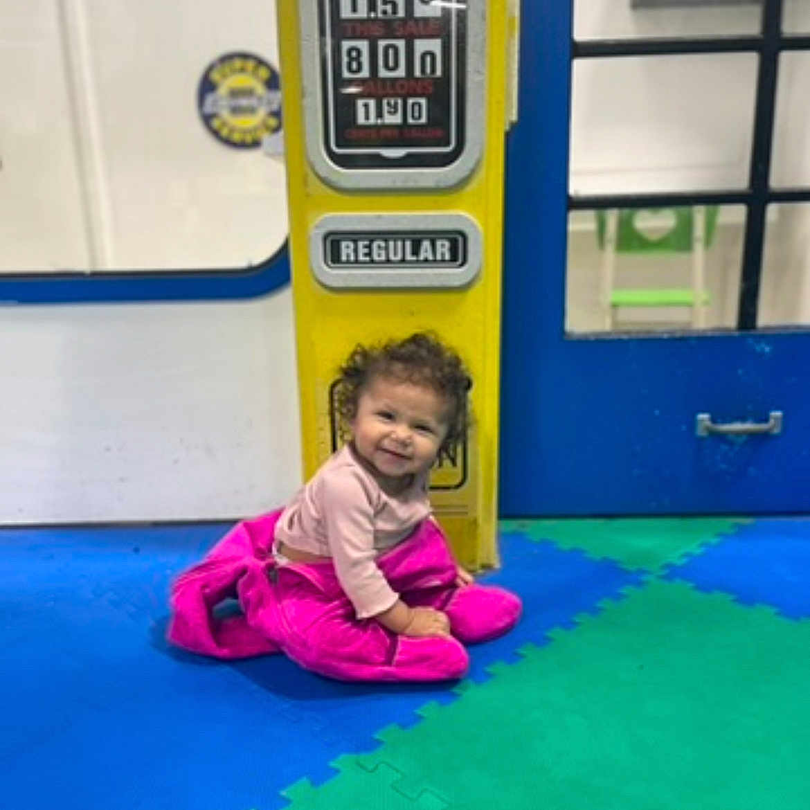 Bryelle joined the competition — help win amazing prizes! blue_floor, child, clothing, curly_hair, door, floor, foam_mats, green_floor, happy, indoor, person, pink_jacket, play_area, portrait, regular_sign, sitting, smiling, toddler, window, yellow_gas_pump