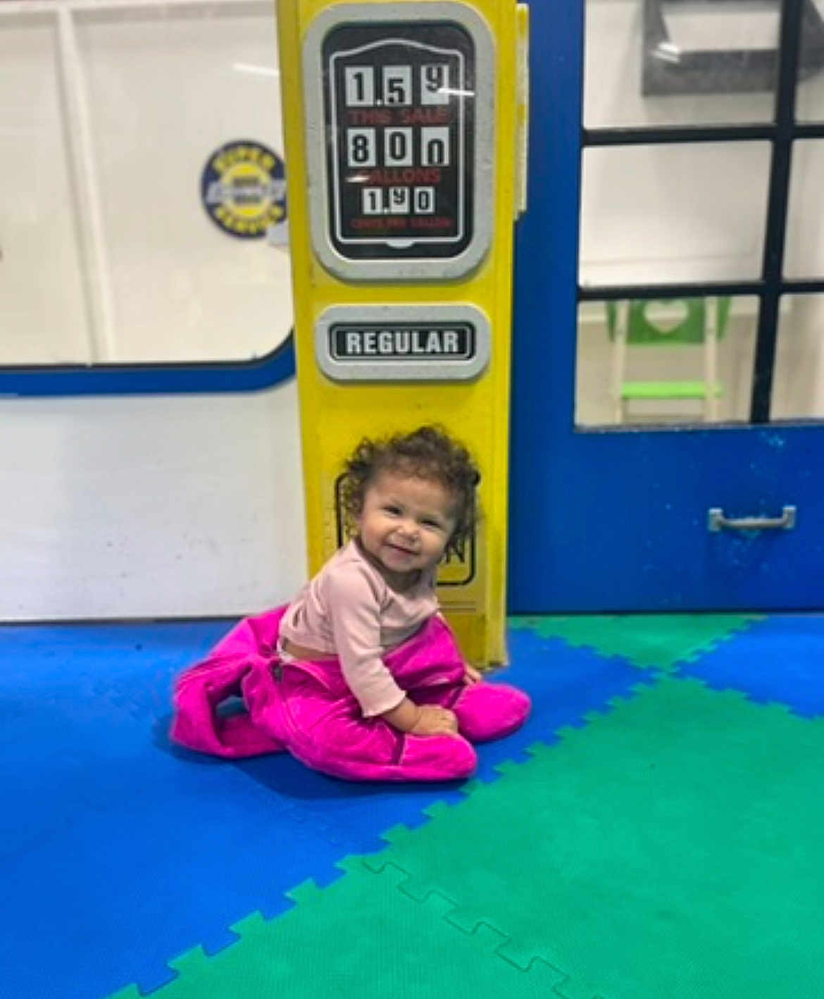 Bryelle joined the competition — help win amazing prizes! toddler, child, smiling, curly_hair, pink_jacket, yellow_gas_pump, regular_sign, foam_mats, blue_floor, green_floor, indoor, play_area, door, window, happy, person, floor, clothing, sitting, portrait