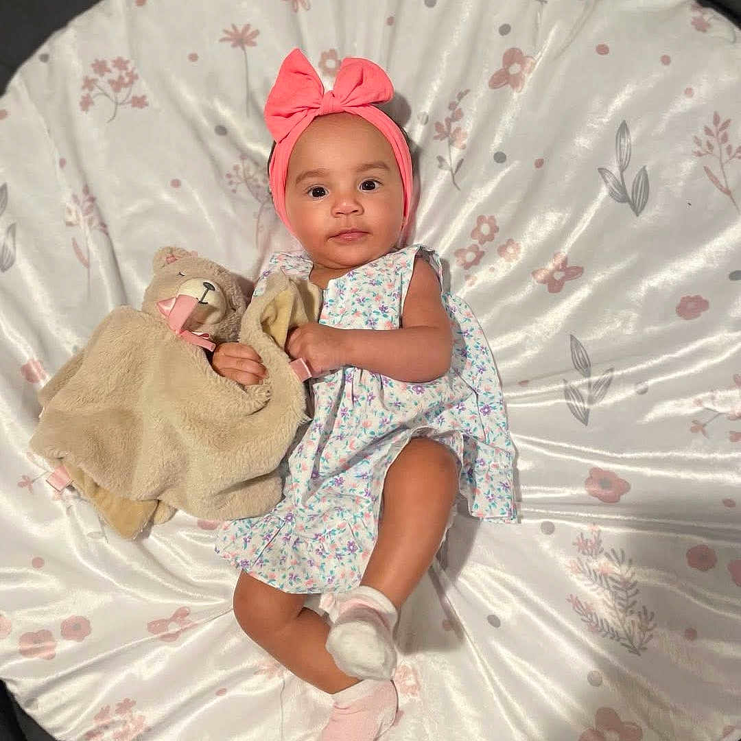 Maci is registered to the contest to win money with this photo: adorable, baby, blanket, bow, child, comfort, cute, dress, floral_pattern, headband, indoors, infant, lying_down, person, pink, plush_toy, sleepy, socks, soft_toy, toddler
