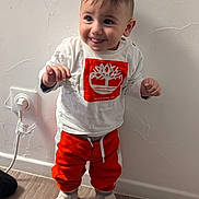 Juan Antonio a rejoint le concours — aidez-le/la à gagner de superbes lots ! child, toddler, baby, smile, standing, white_shirt, orange_pants, sneakers, hands, hair, face, indoor, wall_texture, electrical_outlet, hardwood_floor, portrait, clothing, cute, expression, floor