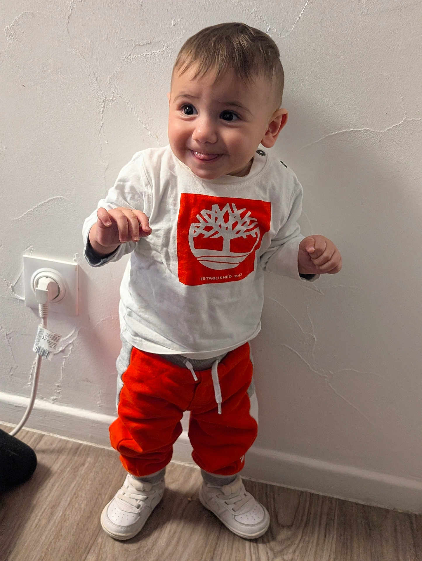 Juan Antonio a rejoint le concours — aidez-le/la à gagner de superbes lots ! child, toddler, baby, smile, standing, white_shirt, orange_pants, sneakers, hands, hair, face, indoor, wall_texture, electrical_outlet, hardwood_floor, portrait, clothing, cute, expression, floor