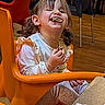 candid, chairs, child, cookie, crumbs, earring, girl, happy, indoor, long_sleeve_shirt, messy_face, orange_chair, patterned_pants, pigtails, portrait, smiling, snack, table, toddler, wooden_floor
