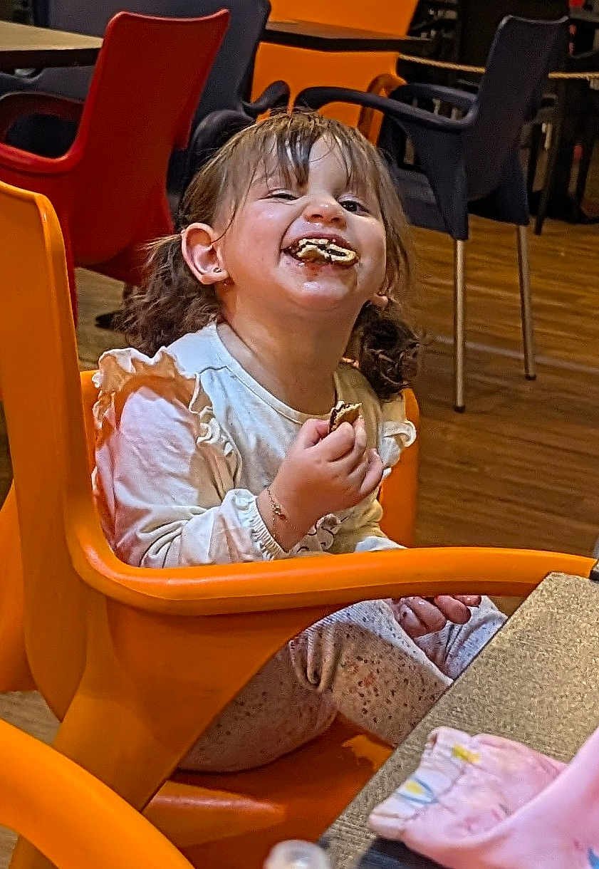 Maria a rejoint le concours — aidez-le/la à gagner de superbes lots ! child, girl, cookie, messy_face, orange_chair, pigtails, smiling, snack, indoor, table, wooden_floor, chairs, portrait, candid, long_sleeve_shirt, patterned_pants, earring, crumbs, happy, toddler
