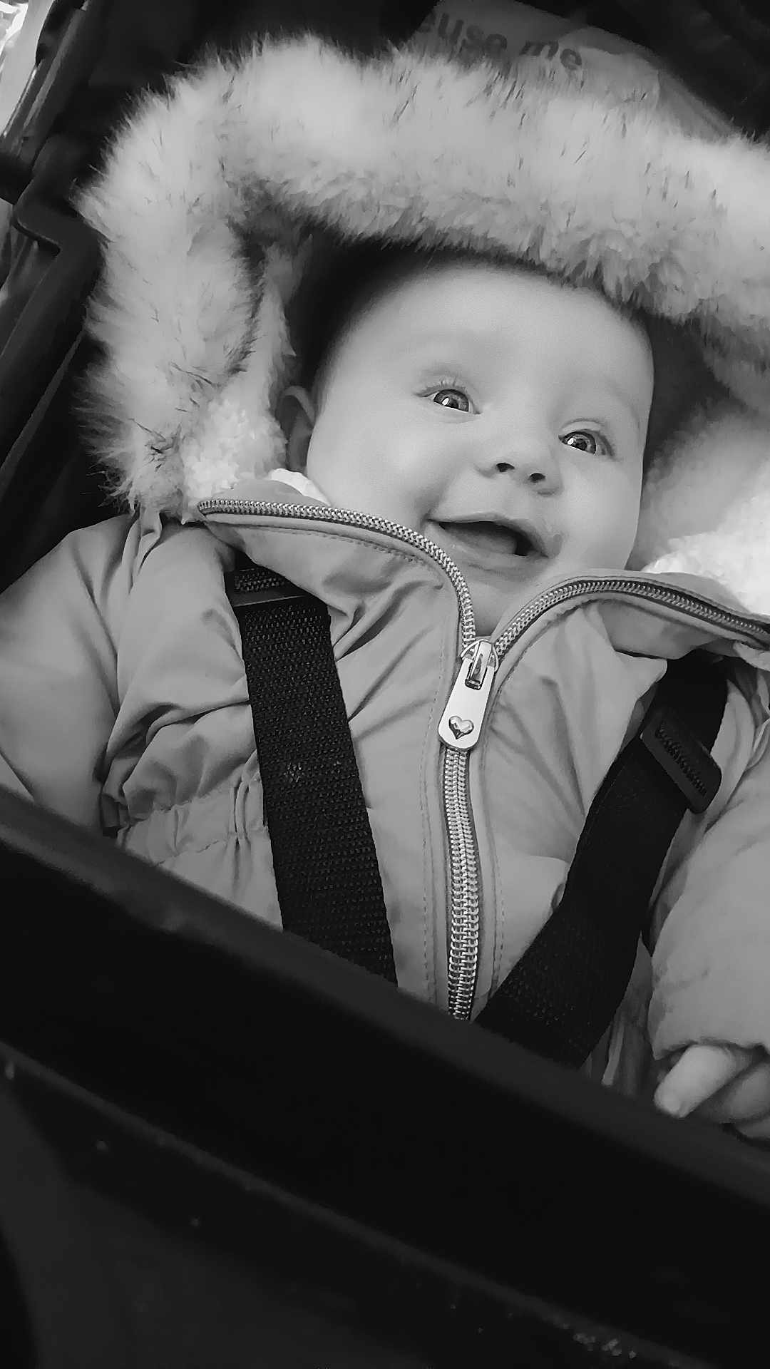 Maisie-May is registered to the contest to win money with this photo: baby, child, smiling, jacket, winter_clothing, fur_hood, stroller, seatbelt, black_and_white, portrait, happy, face, eyes, infant, cute, outdoor, warm_clothing, young_child, expression, close_up