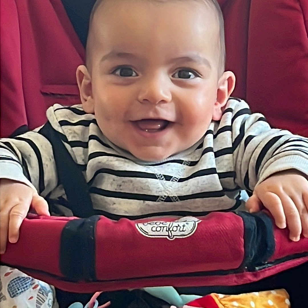 Julian a rejoint le concours — aidez-le/la à gagner de superbes lots ! baby, blanket, bright, child, comfort, cute, face, hand, happy, indoor, infant, person, plush_toys, portrait, red, seated, smiling, striped_clothing, stroller, young