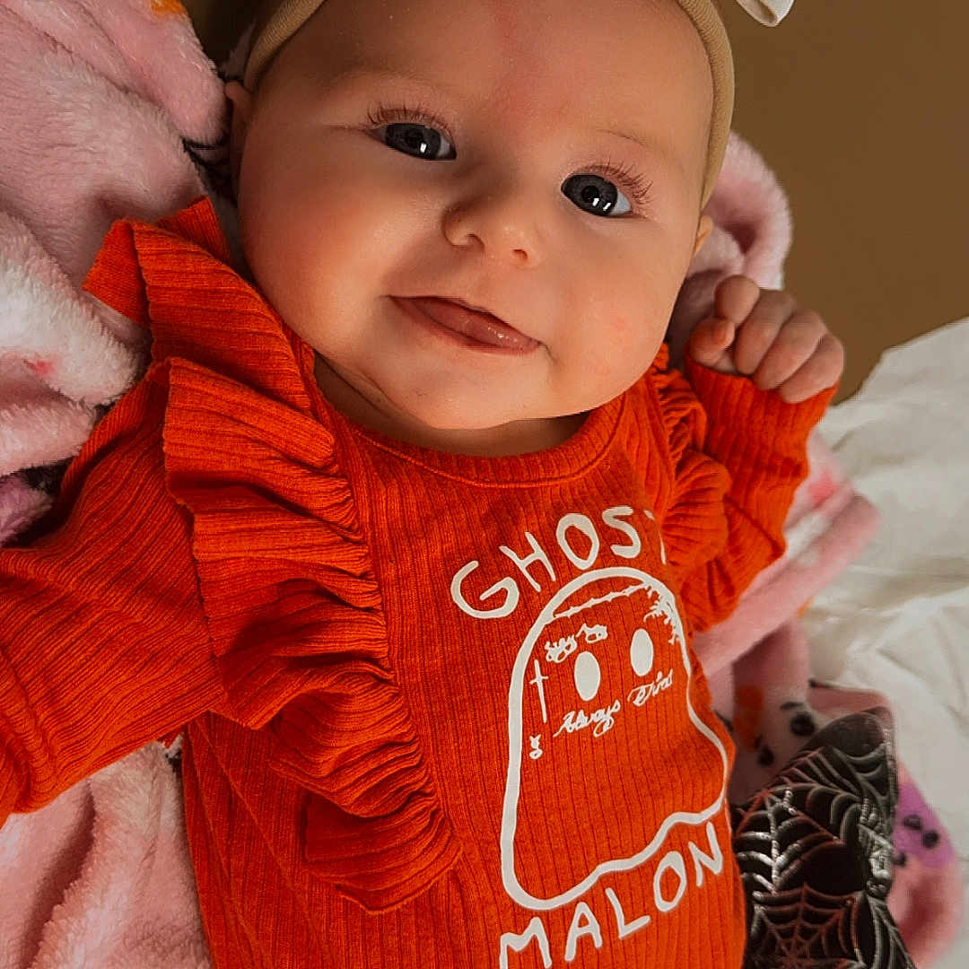 Davina joined the competition — help win amazing prizes! baby, bow, child, clothing, cute, eyes, face, ghost_print, hand, happy, headband, infant, lying_down, orange_clothing, pink_blanket, portrait, ribbon, smiling, soft_texture, toddler