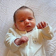 Maxwell is registered to the contest to win money with this photo: baby, newborn, infant, white_outfit, bear_emblem, lying_down, sheet, hands, curly_hair, face, cute, skin, fist, portrait, child, sleepwear, soft_light, indoors, closeup, expression