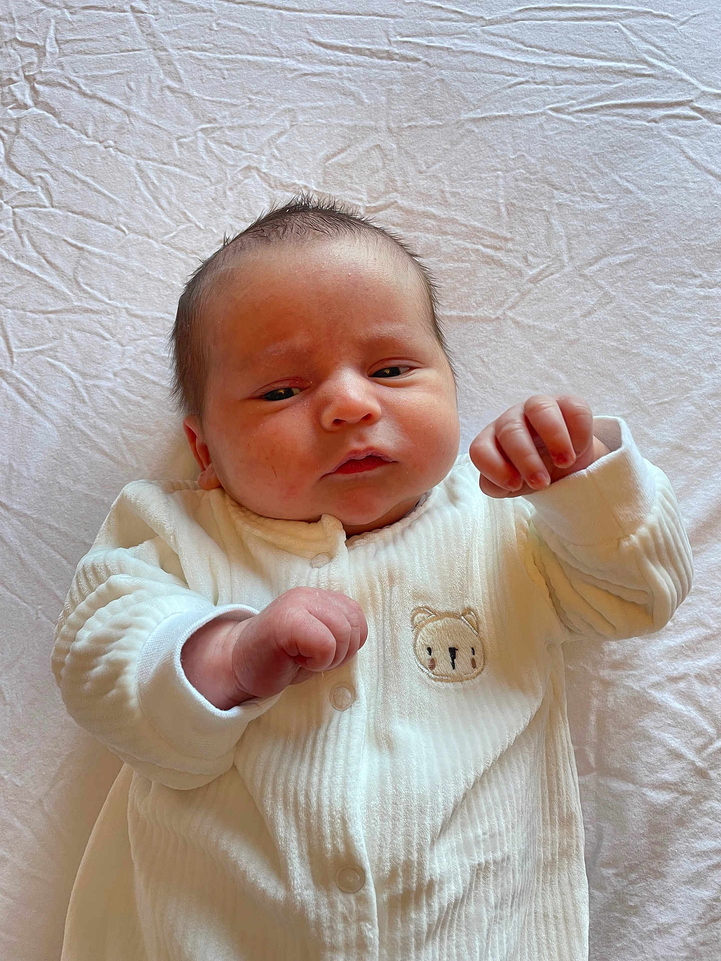 Maxwell is registered to the contest to win money with this photo: baby, newborn, infant, white_outfit, bear_emblem, lying_down, sheet, hands, curly_hair, face, cute, skin, fist, portrait, child, sleepwear, soft_light, indoors, closeup, expression