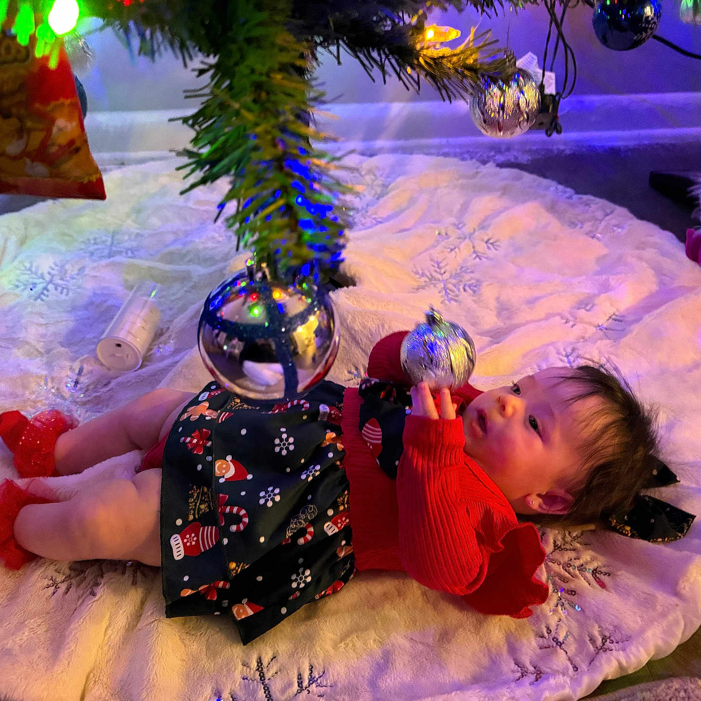 Hanny is registered to the contest to win money with this photo: baby, celebration, child, christmas_tree, curly_hair, decorations, festive, floor, holiday, holiday_season, indoor, lights, ornament, plush, red_clothing, reflection, socks, toy, tree_skirt, unicorn
