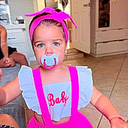 Valentina is registered to the contest to win money with this photo: toddler, child, baby, portrait, pacifier, pink, bow, headband, dress, suspenders, outdoorless, indoors, home, living_room, kitchen, tile_floor, furniture, 成人, sitting, face