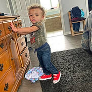Oliver is registered to the contest to win money with this photo: toddler, child, curly_hair, red_sneakers, denim_jeans, tshirt, wooden_dresser, carpet, blanket, indoor, room, doorway, chair, clothes, floor, standing, leaning, casual_clothing, light, home