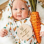 baby, infant, child, blue_eyes, carrot_pattern, carrot_prop, easter_plaque, wooden_sign, hood, onesie, cute, portrait, closeup, soft_blanket, hand, smile, holiday, props, fabric_pattern, woven_decor