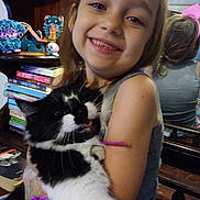 Daisy is registered to the contest to win money with this photo: girl, child, cat, black_and_white_cat, hugging, smiling, portrait, indoor, home, mirror, bookshelf, books, toys, stuffed_animal, gray_shirt, long_hair, pet, animal, cozy, photograph