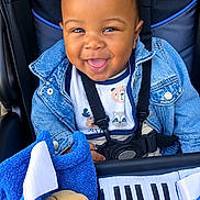 Amari is registered to the contest to win money with this photo: baby, child, smiling, happy, stroller, seated, denim_jacket, bib, toy, stuffed_animal, keyboard_toy, musical_instrument, harness, portrait, indoor, close_up, infant, black_background, blue_clothing, playful