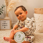 Kaleht Pérez Pardo is registered to the contest to win money with this photo: baby, infant, child, smiling, portrait, sitting, milestone, nine_months, number_nine, sign, indoor, cozy, rug, cushion, pampas_grass, plant, vase, warm_lighting, patterned_clothing, barefoot