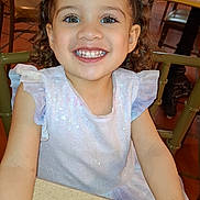 Reychell is registered to the contest to win money with this photo: casual, chair, child, cup, curly_hair, dining, dress, face, girl, glass, happy, indoor, person, restaurant, sitting, smiling, sparkling_eyes, table, wooden_chair, young