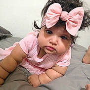 Jane is registered to the contest to win money with this photo: baby, child, infant, portrait, pink_clothing, large_bow, headband, curly_hair, dark_hair, brown_eyes, face, looking_at_camera, lying_down, bed, bedroom, blanket, indoor, close_up, cute, photograph