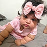 baby, child, infant, portrait, pink_clothing, large_bow, headband, curly_hair, dark_hair, brown_eyes, face, looking_at_camera, lying_down, bed, bedroom, blanket, indoor, close_up, cute, photograph