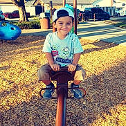 Nicholas is registered to the contest to win money with this photo: child, boy, playground, seesaw, cap, tshirt, toy_story, smile, wood_chips, outdoor, daylight, tree, car, residential, park, play_equipment, footwear, happy, person, recreation