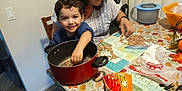 Nicholas is registered to the contest to win money with this photo: beans, chair, child, clothing, cooking, food, hand, happy, indoor, kitchen, package, person, plastic_bag, pot, smiling, snack, table, tablecloth, toddler, woman