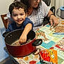 Nicholas is registered to the contest to win money with this photo: toddler, child, woman, pot, beans, table, tablecloth, kitchen, food, snack, cooking, indoor, smiling, happy, person, clothing, plastic_bag, package, chair, hand