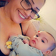 Eli is registered to the contest to win money with this photo: woman, baby, glasses, smile, blanket, indoor, portrait, face, happy, holding, cozy, plant, casual, love, family, comfort, skin, head, expression, warm_light