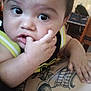 baby, child, finger_in_mouth, tattoo, arm, indoor, curious, closeup, face, person, skin, black_hair, large_eyes, sleeveless_shirt, casual, infant, candid, resting, hand, expression