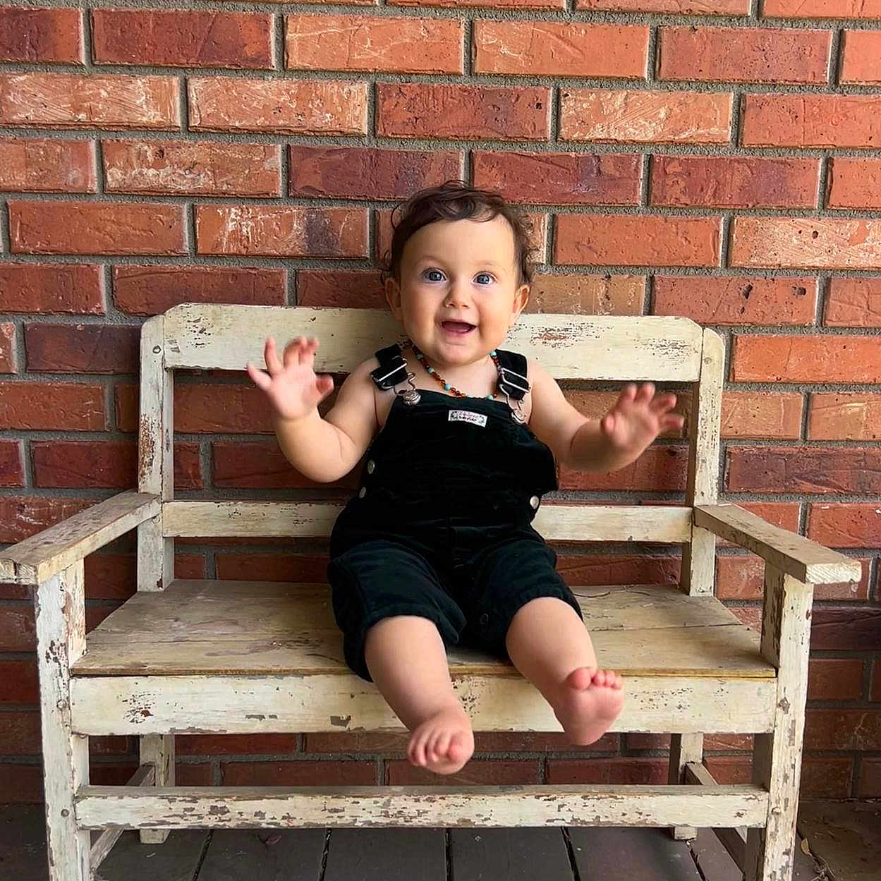 Liv is registered to the contest to win money with this photo: toddler, child, bench, wooden_bench, brick_wall, smiling, happy, barefoot, curly_hair, overalls, outdoor, floor, rustic, portrait, cute, person, sitting, playful, casual, daylight